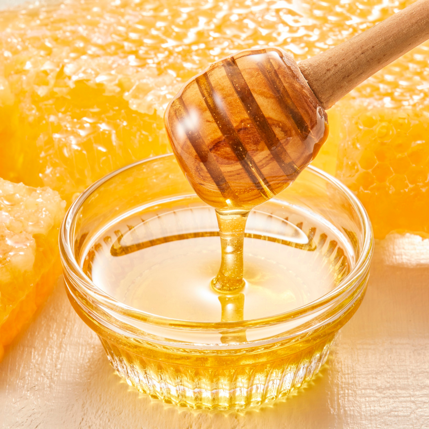 Honeycomb with a wooden honey dipper over a glass bowl filled with golden honey — showcasing Joreen Brands natural unfiltered wild honey in its pure, raw form.